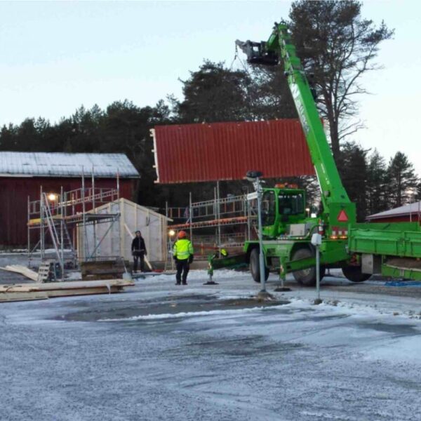 Byggföretaget Umeå fraktar en vägg till ett sopskjul med en stor grön kran. Väggen byggdes i Kronoparken och transporteras till Mariedal i Umeå för montering. Kontakta oss för skräddarsydda bygglösningar i Västerbotten!